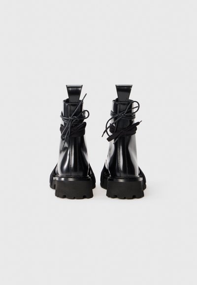Bottines en cuir verni noir avec une semelle épaisse, un bout rond et des lacets en ruban texturé. Comprend des tirettes à l'arrière et des coutures visibles.