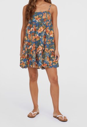 Kvinde iført en blå ærmeløs blomsterkjole med orange blomster og hvide sandaler, stående mod en ensfarvet baggrund.