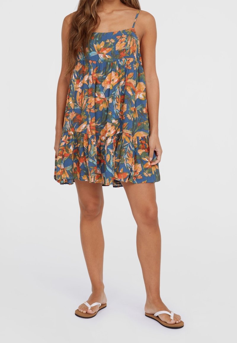 Femme portant une robe fleurie bleue sans manches avec des fleurs orange et des sandales blanches, debout devant un fond uni.