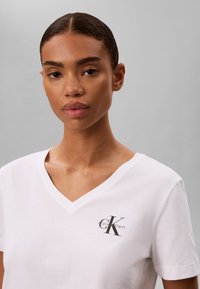 Γυναίκα που φοράει λευκό T-shirt με V-λαιμόκοψη και μικρό μαύρο λογότυπο Calvin Klein στο αριστερό στήθος, σε μονόχρωμο γκρι φόντο.
