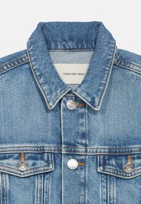 Heleheline teksajakk, millel on terav krae, hõbedast nupud, kaks rinnataskut ja silt, millel on kiri "Calvin Klein Jeans."