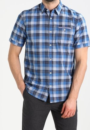 Man in een blauw-grijs geruit overhemd met korte mouwen en zak, staand met één hand in de zak en de andere bij zijn dij.