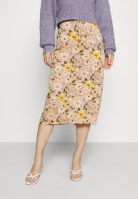 Blommig midi kjol i ljusrosa med gula och gröna mönster; tyget är slätt med en lätt glans. Stylar med vita sandaler med remmar.