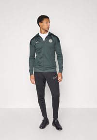 Groene zip-up jas met capuchon, voorzien van een wit Nike-logo en het Chelsea FC-embleem. Zwarte trainingsbroek met zijlogo, gecombineerd met zwarte sneakers.