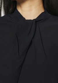 Blouse noire avec un col montant agrémenté d'un nœud, texture de tissu lisse et tombé ample.