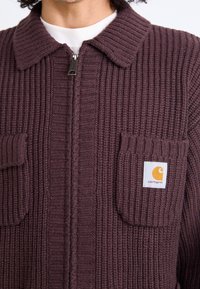 Bordeauxfarbene Strickjacke mit geripptem Muster, Kragen, Frontreißverschluss und zwei Brusttaschen mit dem Carhartt-Logo.