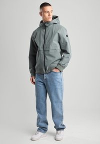 Light grey, lightweight jacket with a hood, black zipper, and two front pockets. Paired with blue jeans and white sneakers.