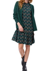 Vestido floral verde con dobladillo con volantes, combinado con un cárdigan de textura verde oscuro y botas negras hasta el tobillo.