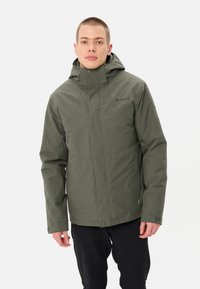 Jeune homme portant une veste d'extérieur à capuche olive et un pantalon noir, se tenant devant un fond clair et uni.