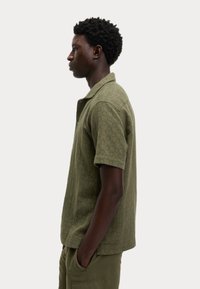Camisa de manga corta verde oliva en tejido texturizado con un sutil patrón geométrico. Cuenta con un collar puntiagudo y un corte relajado.
