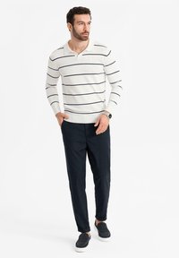 Muž v bílém polo tričku s dlouhým rukávem a černými pruhy, černých kalhotách, černých slip-on botách, s hodinkami na levém zápěstí, stojící s rukama v kapsách.