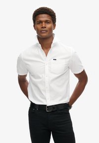 Camisa blanca de manga corta con botones y un bolsillo en el pecho, complementada con un cinturón negro y jeans oscuros. Textura suave, diseño de cuello clásico.