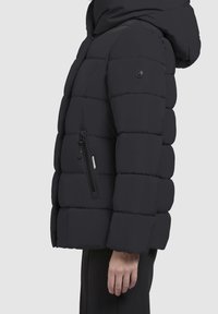 Henkilön sivukuva musta tikattu toppatakki, jossa on vetoketjullinen tasku ja kiinnitetty huppu.