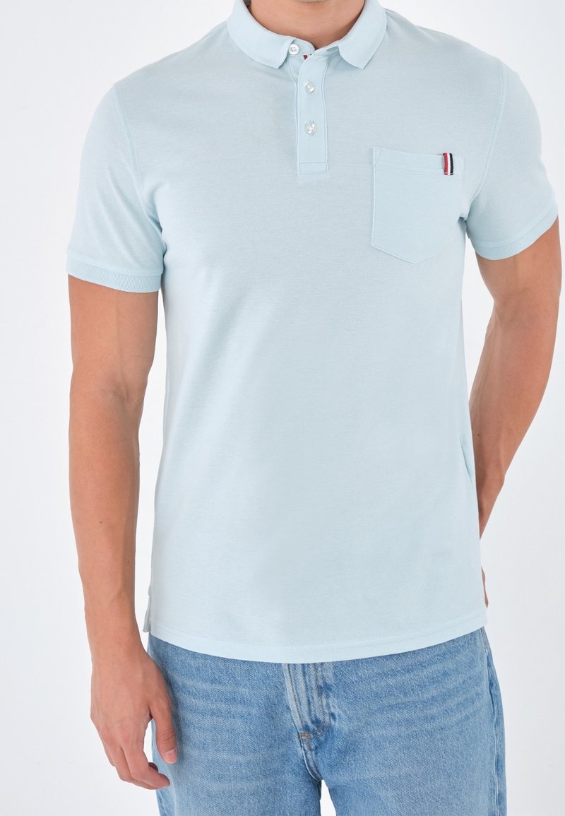 Homme portant un polo bleu clair à manches courtes avec une poche poitrine et des boutons, associé à un jean bleu moyen, debout devant un fond blanc.