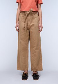 Beige wijde broek met een riem, gemaakt van een gladde katoenstof. Beschikt over zijzakken en zichtbare stikdetails. Zwarte schoenen.