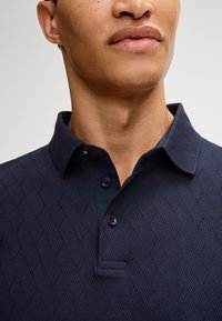 Polo di colore blu navy con un motivo a diamante testurizzato, collettino classico e apertura con tre bottoni. Il materiale sembra essere tessuto e stretch.