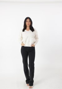Maglione crema a scollo a V con dettagli a motivo cuore dorato, abbinato a jeans neri aderenti. La modella sta in piedi con sicurezza, con le mani nelle tasche.