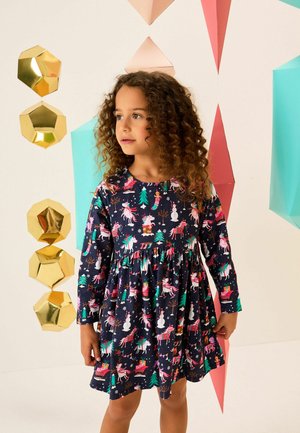 Junges Mädchen mit lockigem Haar, das ein marineblaues Kleid mit Einhorn-, Schneemann- und Weihnachtsbaum-Motiven trägt, steht vor bunten geometrischen Dekorationen.