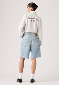 Γυναίκα με μαλλιά σε κότσο, φοράει ανοιχτό γκρι μπουφάν Levi Strauss, ανοιχτό μπλε τζιν σορτς μέχρι το γόνατο, λευκές κάλτσες και μαύρα παπούτσια, στραμμένη προς τα πίσω.