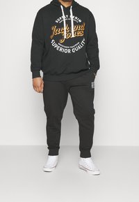 Svart huvtröja med tryckt text i orange och vitt, snörning och ribbade muddar, parat med svarta sweatpants och vita sneakers.