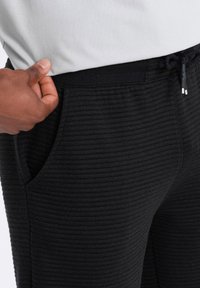 Pantalons noirs avec un design texturé et côtelé, dotés d'une taille élastique à cordon de serrage et de poches latérales. Le tissu semble doux et confortable.