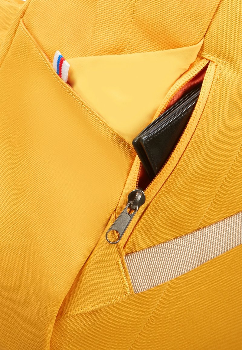 Section d'un sac à dos en tissu jaune avec une poche zippée révélant un portefeuille noir. Présente une bande de tissu horizontale et une texture lisse.