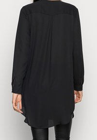 Blusa negra de manga larga con dobladillo redondeado, detalle de pliegues en la parte posterior y puños abotonados. Confeccionada en una tela ligera y suave.