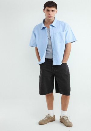 Junger Mann steht mit den Händen in den Taschen, trägt ein hellblaues kurzärmeliges Hemd, ein graues Tanktop, schwarze knielange Shorts, weiße Socken und beige Sneaker.