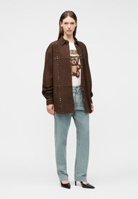 Γυναίκα με καφέ τζάκετ με τρουκς πάνω από λευκό μπλουζάκι με γραφικό αρκουδάκι, ανοιχτό μπλε τζιν και μαύρα ψηλοτάκουνα μυτερά παπούτσια, στέκεται μπροστά σε λευκό φόντο.