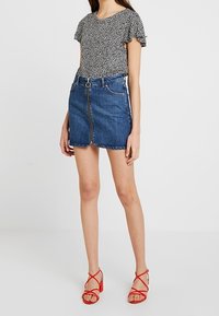 Černobílý top s puntíky a řasenými rukávy, kombinovaný s denimovou mini sukýnkou se středním zipem a tmavým prošitím; červené sandály na vysokém podpatku.