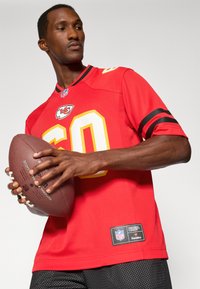 Sarkans NFL krekls ar dzelteniem akcentiem un numuru "60"; īsas piedurknes ar melnām svītrām. Spēlētājs tur futbolu; apakšmalā ir auduma etiķete.