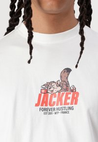 Hvid bomulds t-shirt med grafik af en tegneserie-squirrel med TNT, rød "JACKER" tekst, og "FOREVER HUSTLING" nedenunder i sort.