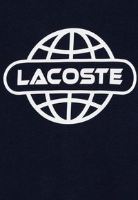 Tela azul marino con un gran diseño de globo blanco y el texto "LACOSTE" en una fuente moderna y negrita centrado debajo.