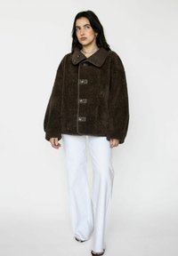Bruine pluche jas met een brede kraag, voorzien van vier knopen en een gladde textuur, gecombineerd met witte flared broek voor contrast.