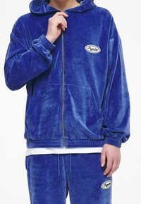 Sudadera con cremallera de terciopelo azul, con un bolsillo frontal, puños acanalados y parche de logo bordado. Combinada con pantalones a juego. Textura suave.