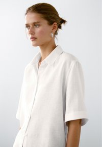 Wit linnen overhemd met korte mouwen en een puntige kraag, gemaakt van licht textuurstof en met knopen aan de voorkant, getoond op een model.