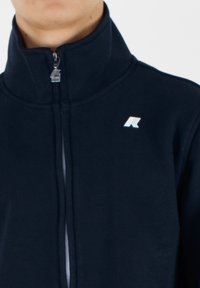 Giacca in pile blu navy con colletto alto, zip frontale completa e logo riflettente sul lato sinistro del petto. Texture morbida e design a taglio dritto.