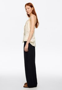Jedwabny top na ramiączkach w kolorze ecru z koronkową lamówką, zestawiony z czarnymi, szerokimi spodniami z wysokim stanem oraz brązowymi wsuwanymi butami.