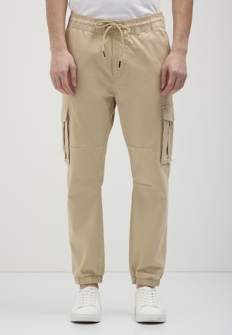 Pantaloni cargo beige con polsini e vita elasticizzati, dotati di tasche laterali e vestibilità comoda. Il materiale sembra leggero e liscio.