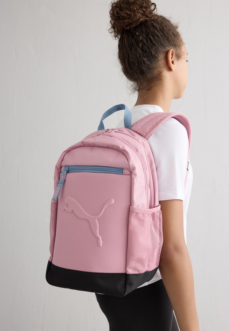 Sac à dos en tissu rose avec une texture lisse, dotée d'une fermeture éclair bleue, d'une poche latérale en mesh et d'un fond noir. Logo bien visible.