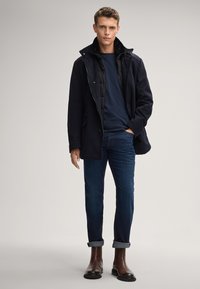 Marineblauer Mantel mit einem Kragen aus Kunstfell über einem dunkelblauen Hemd, kombiniert mit hochgekrempelten Jeans und braunen Stiefeletten. Elegantes und modernes Design.