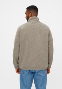 Pulloverjakke i lys grå fleece med høj krave, raglanærmer og en struktureret overflade, kombineret med blå jeans.