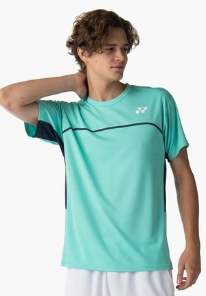 Giovane uomo che indossa una maglietta sportiva turchese chiaro con accenti blu scuro e pantaloncini bianchi, in posa con la mano dietro il collo, guardando verso destra.