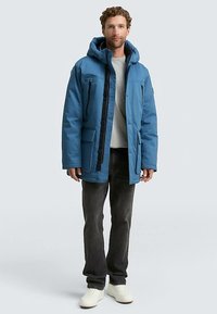 Zila izolēta jaka ar kapuci, priekšpusē rāvējslēdzējs, aizdare ar pogām, vairākas kabatas un melni akcenti. Valkāta pāri gaišam džemperim.