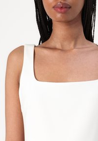 Vit top med kvadratisk halsringning som har breda axelband och en mjuk textur, vilket framhäver halsringningen och axelpartiet.