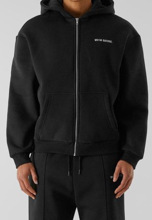 Felpa nera con zip e piccolo testo bianco "9N1M SENSE." sul petto, indossata da una persona con le mani rilassate ai lati, abbinata a pantaloni neri coordinati.