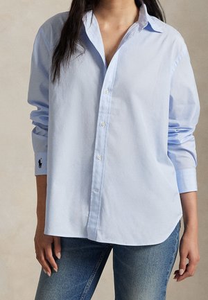 Kvinde iført en lyseblå oversized skjorte med opkrogede ærmer og blå jeans, stående med afslappet kropssprog.
