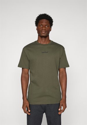 Man standing facing forward wearing olive green short-sleeve t-shirt with small central logo and dark gray pants on plain background.