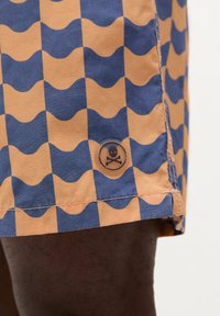 Shorts de bain avec un motif de vagues orange et bleu, ornés d'un détail en bouton illustrant un crâne et des os croisés sur l'ourlet.
