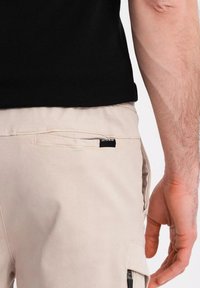Pantalons cargo beiges avec une poche latérale, texture du tissu visible. Étiquette logo noire sur la poche. Portés avec un haut noir, montrant une main sur la taille.
